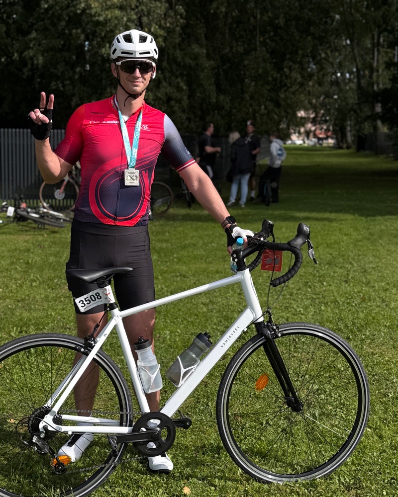 Vsevolod standing in a park beside a white road bike in a red cycling jersey, white helmet and finisher medal, making a peace sign